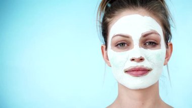 Yüzünde beyaz kuru çamur maskesi olan genç bir kadın, maviliğe karşı. Genç kız yağlı cildiyle ilgileniyor, gözenekleri temizliyor. Güzellik tedavisi. Cilt bakımı.