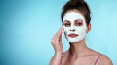 Yüzünde beyaz kurumuş çamur maskesi olan genç bir kadın pamuklu çubukla maviden temizleniyor. Yağlı ciltle ilgilenen genç bir kız. Güzellik tedavisi. Cilt bakımı.