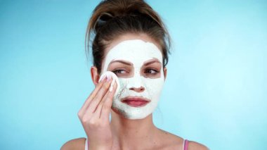 Yüzünde beyaz kurumuş çamur maskesi olan genç bir kadın pamuklu çubukla maviden temizleniyor. Yağlı ciltle ilgilenen genç bir kız. Güzellik tedavisi. Cilt bakımı.