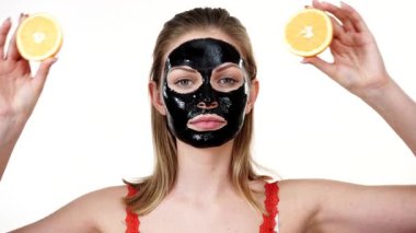 Yüzünde karbonhidrat maskesi olan genç bir kadın elinde turuncu meyve yarımları, gözleri kaplayan beyaz bir kadın. Güzellik tedavisi. Cilt bakımı.