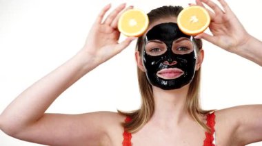 Yüzünde karbonhidrat maskesi olan genç bir kadın elinde turuncu meyve yarımları, gözleri kaplayan beyaz bir kadın. Güzellik tedavisi. Cilt bakımı.