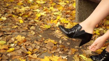 Sonbahar parkında dinlenen bir kadın. Kadın bankta oturuyor, ayaklarındaki siyah deri ayakkabıyı giyip fermuarını çekiyor.
