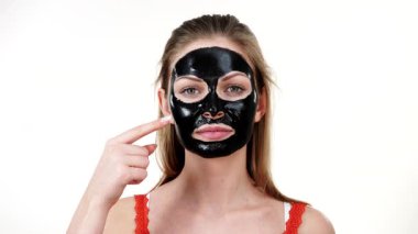 Karbonhidrat maskesi takmış genç bir kadın. Yüzünün, beyazının farklı bölgelerine parmağıyla işaret ediyor. Genç kız siyah gözenekleri temizliyor. Güzellik tedavisi. Cilt bakımı.