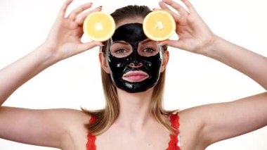 Yüzünde karbonhidrat maskesi olan genç bir kadın elinde turuncu meyve yarımları, gözleri kaplayan beyaz bir kadın. Güzellik tedavisi. Cilt bakımı.