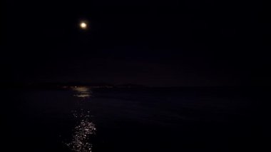 Gece dolunayda, kara gökyüzünde, kıyı bölgesinde. Ay ışığı suya yansıyor.