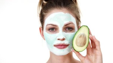 Yüzünde beyaz yeşil çamur maskesi olan genç bir kadın avokado meyvesi tutuyor. Genç kız cildine bakıyor, gözenekleri temizliyor. Güzellik tedavisi.