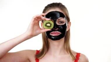 Yüzünde karbonhidrat maskesi olan genç bir kadın elinde kivi meyve yarımları, gözleri kaplayan beyaz bir kadın. Güzellik tedavisi. Cilt bakımı.