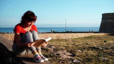 Olgun turist kadın kitapla sahilde dinleniyor, temiz havanın tadını çıkarıyor. Tatillerde kitap okumak. Dinlenme zamanı..