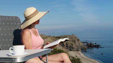 Kitap ve kahve fincanıyla sahilde dinlenen olgun bir turist kadın, uçurumdan deniz manzarasının tadını çıkarıyor. Tatillerde kitap okumak. Dinlenme zamanı..