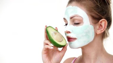 Yüzünde beyaz yeşil çamur maskesi olan genç bir kadın avokado meyvesi tutuyor. Genç kız cildine bakıyor, gözenekleri temizliyor. Güzellik tedavisi.