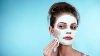 Yüzünde beyaz kurumuş çamur maskesi olan genç bir kadın pamuklu çubukla maviden temizleniyor. Yağlı ciltle ilgilenen genç bir kız. Güzellik tedavisi. Cilt bakımı.