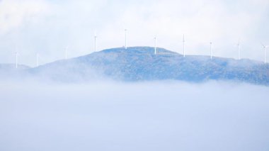 Rüzgâr türbinleri sisli arazide tepede bir çiftlikte. Yel değirmeni sabah bulutlarla kaplıydı. Yeşil ekolojik enerji üretimi.