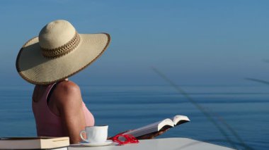 Kitap ve kahve fincanıyla sahilde dinlenen olgun bir turist kadın, uçurumdan deniz manzarasının tadını çıkarıyor. Tatillerde kitap okumak. Dinlenme zamanı..