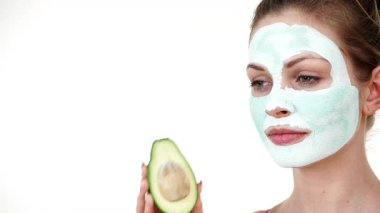 Yüzünde beyaz yeşil çamur maskesi olan genç bir kadın avokado meyvesi tutuyor. Genç kız cildine bakıyor, gözenekleri temizliyor. Güzellik tedavisi.