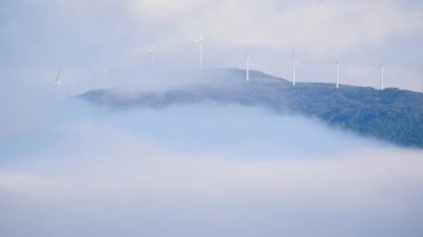Rüzgâr türbinleri sisli arazide tepede bir çiftlikte. Yel değirmeni sabah bulutlarla kaplıydı. Yeşil ekolojik enerji üretimi.