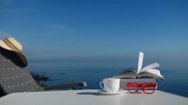 Tatillerde kitap okumak. Kahve fincanı ve kitaplar açık havada mavi deniz suyuna karşı. Kitap sayfaları rüzgarda çırpınıyor.