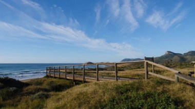Sahil manzarası, denize giden ahşap yolu olan deniz kıyısı, dalgalar ağır çekimde hareket ediyor. Cala Magre Sahili, Calique Bölgesel Parkı, Murcia Bölgesi İspanya.