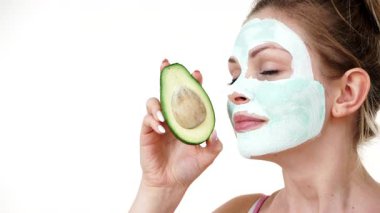 Yüzünde beyaz yeşil çamur maskesi olan genç bir kadın avokado meyvesi tutuyor. Genç kız cildine bakıyor, gözenekleri temizliyor. Güzellik tedavisi.