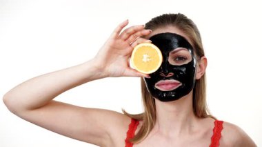 Yüzünde karbonhidrat maskesi olan genç bir kadın portakal meyvesi tutuyor, gözü kapalı, beyaz üzerinde. Güzellik tedavisi. Cilt bakımı.