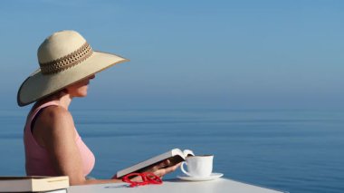 Kitap ve kahve fincanıyla sahilde dinlenen olgun bir turist kadın, uçurumdan deniz manzarasının tadını çıkarıyor. Tatillerde kitap okumak. Dinlenme zamanı..