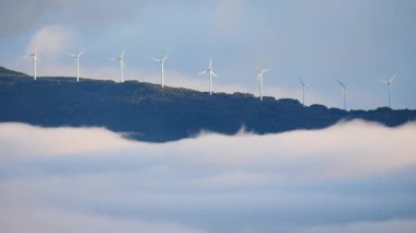 Sabahları bulutların üstündeki tepede rüzgâr türbinleri çiftliği. Koruma ve sürdürülebilir enerji kavramı.