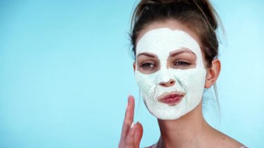Yüzünde beyaz kuru çamur maskesi olan genç bir kadın, maviliğe karşı. Genç kız yağlı cildiyle ilgileniyor, gözenekleri temizliyor. Güzellik tedavisi. Cilt bakımı.