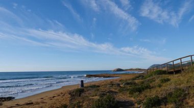 Sahil manzarası, denize giden ahşap yolu olan deniz kıyısı, dalgalar ağır çekimde hareket ediyor. Cala Magre Sahili, Calique Bölgesel Parkı, Murcia Bölgesi İspanya.