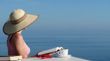 Kitap ve kahve fincanıyla sahilde dinlenen olgun bir turist kadın, uçurumdan deniz manzarasının tadını çıkarıyor. Tatillerde kitap okumak. Dinlenme zamanı..