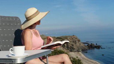 Kitap ve kahve fincanıyla sahilde dinlenen olgun bir turist kadın, uçurumdan deniz manzarasının tadını çıkarıyor. Tatillerde kitap okumak. Dinlenme zamanı..