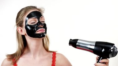 Yüzünde karbonhidrat maskesi olan genç bir kadın. Saç kurutma makinesini kullanarak, beyazın üzerinde kurutmak için kozmetik ürünlerini kurutan bir kız. Güzellik tedavisi. Cilt bakımı.