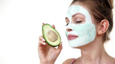 Yüzünde beyaz yeşil çamur maskesi olan genç bir kadın avokado meyvesi tutuyor. Genç kız cildine bakıyor, gözenekleri temizliyor. Güzellik tedavisi.