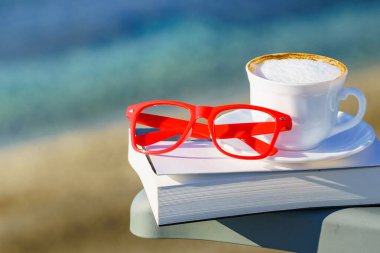 Ruhsal iyileşme, pozitif terapi. Sahile karşı kahve fincanı, kitap ve kırmızı bardaklar. Tatilde okumak. Tatillerde rahatla. Zihinsel sağlık sorunları.