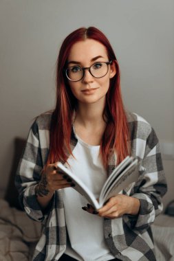 Ekose gömlekli ve gözlüklü kızıl saçlı bir kız elinde bir kitap tutuyor.