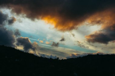 Karamsar bulutlar ve turuncu röfleler ile gün batımı gökyüzü ormanlı tepeler üzerine dramatik ışık saçıyor.