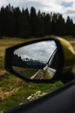 Dağ manzarası araba yan aynasına yansıyor ve yaratıcı bir seyahat perspektifi var. Kabarık bulutlar ve orman arka planı.