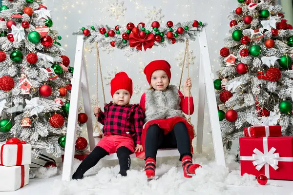 Süslü bir Noel odasında salıncakta oturan iki mutlu kız kardeşin portreleri. Kırmızı şenlik kıyafetleri içinde. Mutlu çocuklar Noel 'de.