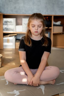 Gözleri kapalı küçük neşeli kız evde yoga pozu veriyor.