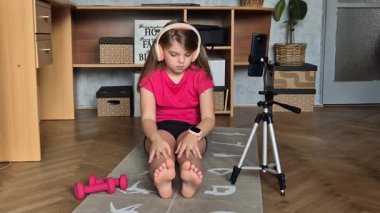 Yoga, evde online spor. Kulaklıklı bir çocuk evde yoga minderinde tren kullanıyor, akıllı telefondan internetten eğitim görüyor. Çocuk sağlığı hizmetleri.