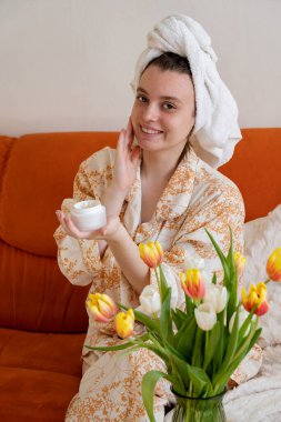 Evde günlük hayatta cilt bakım kremi kullanan ev elbiseli genç bir kız. Cilt bakım ürünü, nemlendirici ya da losyon. Evde yüz bakımı..