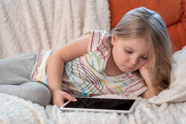 Elinde tablet bilgisayarla turuncu bir kanepeye uzanmış evde ders çalışan küçük bir kız. Online dersler. Uygulamaları kullanan çocuk, internette gezinme bilgileri.