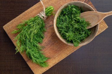 Kahverengi mutfak tahtasındaki ahşap bir tabakta taze dereotunun yanında doğranmış taze dereotu. Donmaya hazırlanıyorum. Yemek pişirmek için aromatik bitkiler kavramı.