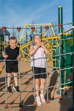Neşeli oğlan ve kız yazın oyun parkında ip merdivene tırmanıyorlar. Spor ve sağlıklı çocuk yaşam tarzı. Açık hava oyunları.