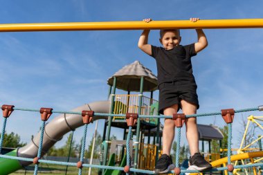 Neşeli küçük çocuk yazın oyun alanında yatay çubuklara ve ip merdivene tırmanıyor. Spor ve sağlıklı çocuk yaşam tarzı. Açık hava oyunları.