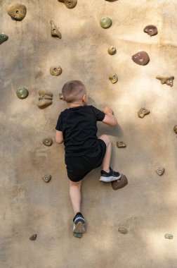 Koyu renk elbiseli küçük çocuk yazın spor sahasında yapay bir kaya duvarına tırmanıyor. Arkadan bak. Okulda beden eğitimi. Çocuk sporu ve sağlıklı bir yaşam tarzı..