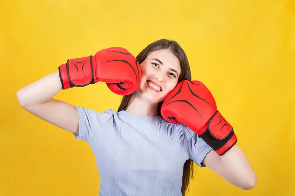 Positive and funny woman with red boxing gloves keeps fists to her cheeks. Portrait of confident girl smiling to camera isolated on yellow background