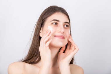 Güzel bir kadın yanaklarına nemlendirici yüz kremi sürüyor ve yan tarafa bakıyor. Güzel kız beyaz arka planda izole edilmiş nemlendirici sürüyor. Cilt bakımı, kozmetik, güzellik kaplıcası konsepti.