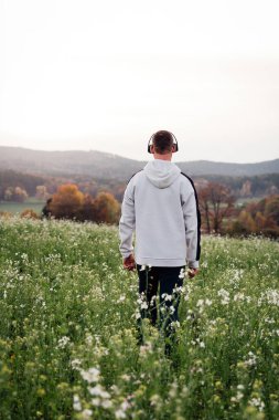 Kapüşonlu ve pantolonlu genç beyaz adam tarlada yürüyor ve kulaklıkla müzik dinliyor. Sonbahar manzarası 