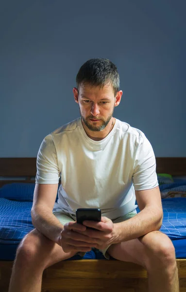 Young caucasian man sitting on bed and using smartphone before going sleep. Negative effect on sleep