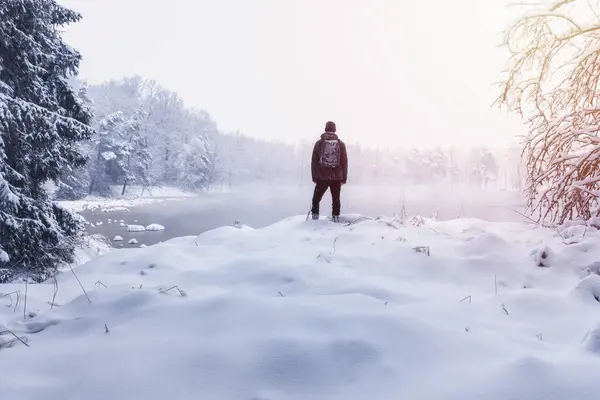 Young man in jacket standing on pond shore with fresh snow in winter storm. Czech landscape background