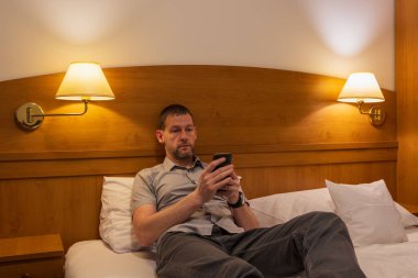 Bir adam otel odasında bir yatakta uzanıyor ve çalışıyor, cep telefonunda rahatlıyor, internette arama yapıyor.